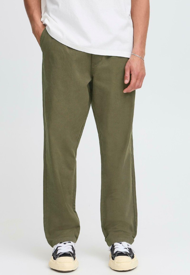 Man wearing olive green pants, black and white sneakers, and a white shirt with one hand in pocket against plain background.