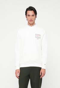 Ung mand iført en hvid sweatshirt med et lille logo på forsiden og mørkegrønne bukser, stående foran en ensfarvet hvid baggrund.