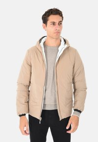 Uomo che indossa un giubbotto imbottito beige con cappuccio sopra un maglione grigio chiaro e pantaloni neri, in piedi contro uno sfondo chiaro uniforme.