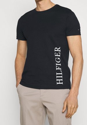 Mężczyzna ubrany w czarną koszulkę z krótkim rękawem z napisem "HILFIGER" pionowymi białymi literami oraz beżowe spodnie, ręce swobodnie opuszczone wzdłuż ciała.