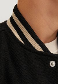Svart jackkrage med en ribbad trim i beige och svart, med en detalj av silverfäste. Mjuk, strukturerad tyg.