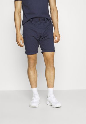 Granatowe bawełniane szorty z podwiniętymi nogawkami, przednimi kieszeniami oraz brązowym zapięciem na guzik. Noszone z dopasowaną górą i białymi trampkami.