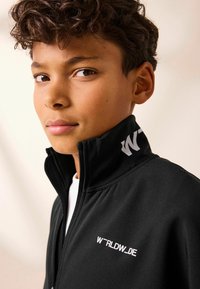 Schwarze Zip-Jacke mit hohem Kragen, mit weißem Logo-Text auf der Brust und dem Kragen. Glatte, matte Textur mit strukturierten Nähten.