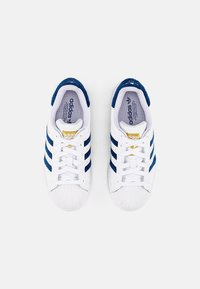 White sneakers with navy blue stripes, featuring a textured leather upper, rubber sole, and a gold logo on the tongue. Flat laces and shell toe.