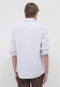 Giovane visto di spalle che indossa una camicia a maniche lunghe di un leggero viola con le maniche arrotolate e pantaloni marrone scuro su uno sfondo semplice.