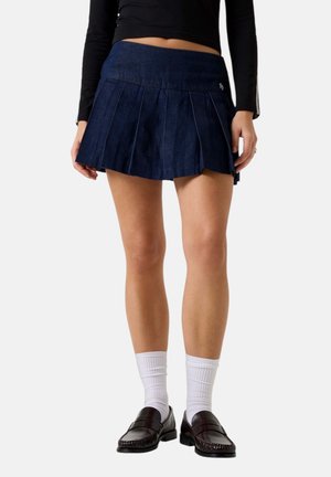 Donna che indossa una minigonna plissettata blu navy, calzini bianchi fino al polpaccio, mocassini neri e una maglia nera a maniche lunghe, in piedi davanti a uno sfondo neutro.