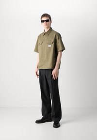 Olive green short-sleeve shirt with two chest pockets, paired with black wide-leg trousers and black shoes, model wearing black sunglasses.