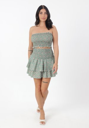 Mujer de pie con un top verde floral sin tirantes con fruncidos y falda a juego en capas con sandalias blancas de tacón sobre un fondo blanco.