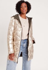 Manteau matelassé doré métallisé avec capuche, design en piqué, et fermeture éclair à l'avant. Porté sur un haut beige clair, associé à un jean large bleu.