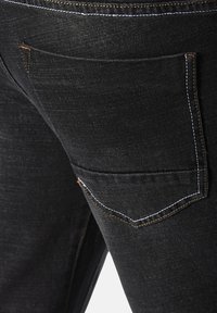 Nahaufnahme einer schwarzen Jeanshose mit einer Gesäßtasche auf der rechten Seite, die mit weißem und braunem Faden abgesteppt ist.