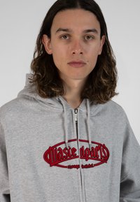 Hoodie gris zippé en tissu mélangé coton, avec logo rouge brodé sur la poitrine. Comprend une capuche avec cordon de serrage et une fermeture éclair argentée.