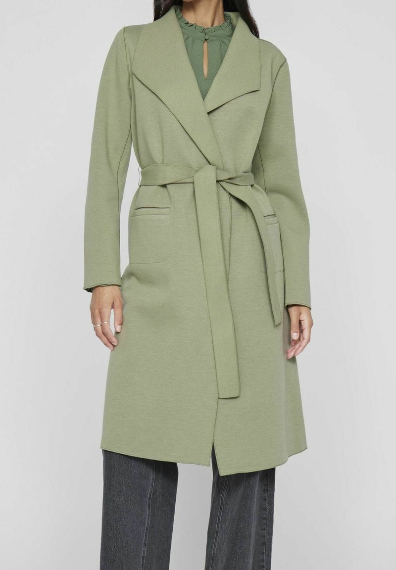 Femme portant un long manteau ceinturé vert clair sur un haut vert, associée à un pantalon gris foncé à jambe droite, debout devant un fond uni.