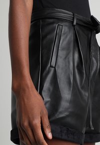 Mustat nahkashortsit, joissa on korkea vyötärö, etumallit, sivutaskut ja pehmeä musta tekoturkisreuna helmassa. Mukana on solkivyön yksityiskohta.