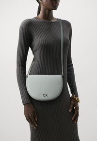 Borsa a spalla in pelle verde chiaro con forma curvata, caratterizzata dal logo "CK" in argento. Indossata con un vestito grigio aderente e a coste.