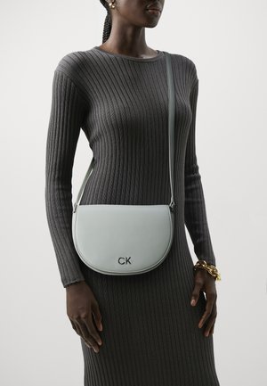 Borsa a spalla in pelle verde chiaro con forma curvata, caratterizzata dal logo "CK" in argento. Indossata con un vestito grigio aderente e a coste.