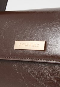 Brown faux leather handbag with a textured surface. Features a gold decorative plaque with "ANNA FIELD" embossed. Stitched edges visible.