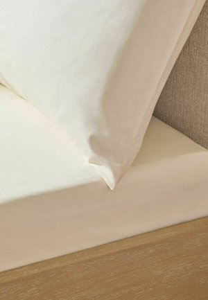Kissen in Cremefarbe und Spannbettlaken auf einem Holzbettgestell mit einem beigen Stoff-Kopfteil.