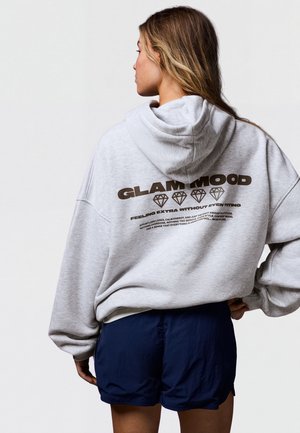 Femme aux longs cheveux portant un sweat à capuche gris avec le texte "GLAM MOOD" et des graphiques de diamants dans le dos, associée à un short bleu foncé, debout avec la main sur la hanche.