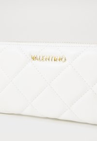 Bela prešitna usnjena denarnica z zlatim zadrgo in zlatimi črkami "VALENTINO" na sprednji strani.