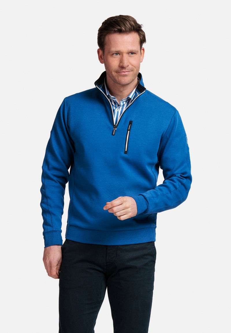 Blauwe sweatshirt met een halve rits, geribde kraag en lange mouwen. Voorzien van een borstzak met een zwarte rits, gecombineerd met donkere broek.