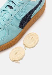 Zapatillas Puma de gamuza azul claro, con acentos en azul oscuro, suela de goma y dos cintas de crema colocadas al lado del zapato.