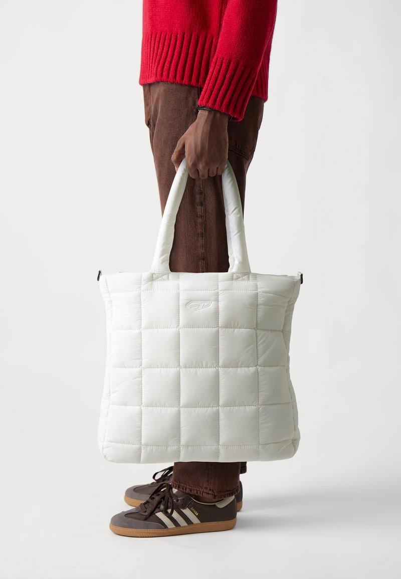 Sac en toile matelassé blanc avec texture rembourrée, doubles poignées et logo discret à l'avant, présenté aux côtés d'une personne en tenue décontractée.