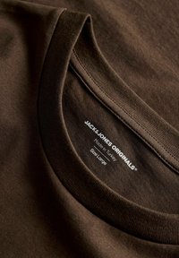 Κοντινό πλάνο στον καφέ γιακά T-shirt με λαιμόκοψη τύπου crew, που δείχνει τυπωμένη ετικέτα με τη μάρκα Jack & Jones Originals, κατασκευασμένο στην Τουρκία, μέγεθος μεγάλο.
