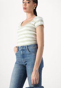 Ανοιχτό πράσινο και λευκό ριγωτό τοπ κοντομάνικο, με ανοιχτό λαιμό, συνδυασμένο με ψηλόμεσα μπλε τζιν. Η υφή φαίνεται ραμμένη.
