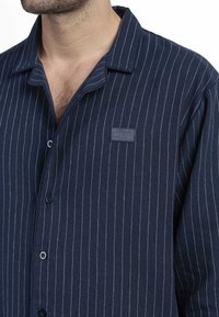 Camisa de botones a rayas marinas hecha de tela suave; presenta rayas verticales blancas, un parche con logo en el pecho y un diseño de cuello clásico.