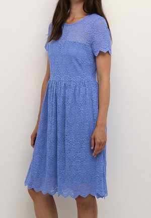 Kvinde iført en knælængde, kortærmet blå blondekjole med blomsterprint og bølget kant, stående foran en ensfarvet baggrund.