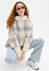 Fleece pullover in cream and gray check pattern, featuring a high collar, zip closure, and puffy sleeves, paired with light blue jeans and sneakers.