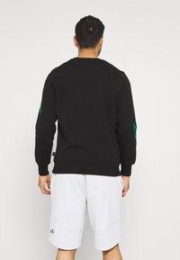 Czarny sweatshirt z zielonymi akcentami graficznymi na rękawach, z okrągłym dekoltem i ściągaczami. Noszony z jasnoszarymi szortami.