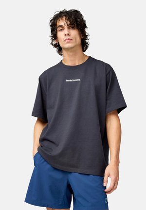 Vīrietis ar cirtainiem matiem valkā vaļīgu melnu "Brooks Running" T-kreklu un zilas sporta šortus, stāvot ar vienu roku kabatā.