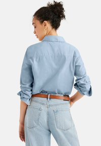 Camicia azzurro chiaro con maniche arrotolate e colletto, abbinata a jeans in denim chiaro e una cintura in pelle marrone.