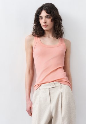 Γυναίκα με σγουρά μαλλιά που φοράει μπεζ τοπάκι χωρίς μανίκια και μπεζ παντελόνι με ψηλή μέση, στέκεται μπροστά σε λευκό, απλό τοίχο.