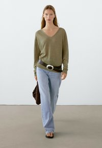 Olivgrüner V-Ausschnitt-Pullover, lockere Passform, kombiniert mit hellblauen weitem Jeans. Schwarzer Gürtel mit silberner Schnalle und brauner strukturierter Tasche. Flache Sandalen.
