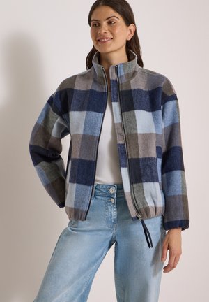 Femme souriante portant une veste zippée à carreaux bleus et gris avec un col montant et un jean bleu clair.