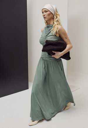 Moteris dėvi rankovių neturintį žalsvai mėtininės spalvos maxi suknelę, baltą galvos juostą, laiko tamsią sulankstytą striukę, vaikšto minimalistiniame patalpų interjere.