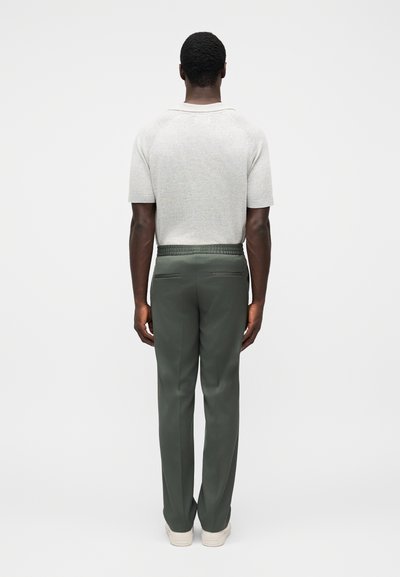 Ανοιχτό γκρι πλεκτό πουλόβερ με κοντά μανίκια, πράσινο καλοραμμένο παντελόνι με ελαστική μέση και εκρού αθλητικά παπούτσια· εμφανίζεται από πίσω.