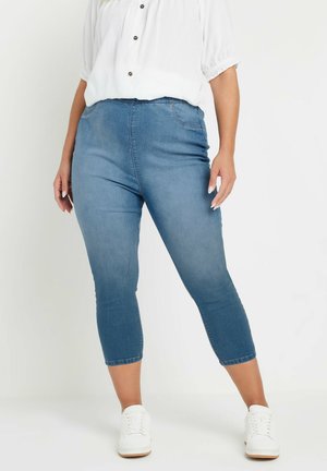 Kvinde iført blå cropped skinny jeans, hvid knappet bluse og hvide sneakers, stående mod en ensfarvet lys baggrund.