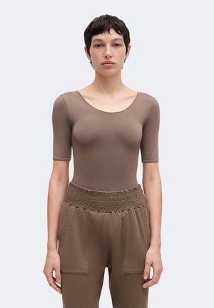 Frau mit kurzem, dunklem Haar trägt ein enganliegendes taupefarbenes Kurzarm-Oberteil und eine hoch taillierte braune Hose, stehend vor einem schlichten, hellen Hintergrund.