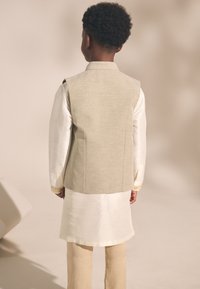 Tenue traditionnelle pour garçons comprenant un gilet beige clair à finition texturée, une chemise blanche et un pantalon couleur crème. Vue de dos.