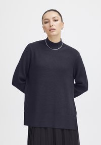 Dunkelblauer, gestrickter Rollkragenpullover mit geripptem Muster, �überschnittenen Schultern und lockerer Passform, kombiniert mit einem fließenden schwarzen Rock.