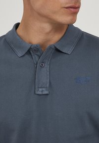 Mørkeblå poloshirt med strukturert stoff, to knapper og en liten brodert logo i samme nyanse nær kragen.