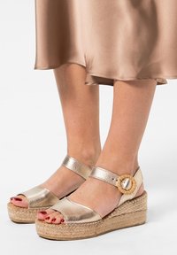 Gouden metallic platform sandalen met open tenen, geweven jute zolen en een ronde sluiting als accent op de enkelband. Gladde textuur.
