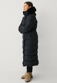 Lange zwarte puffercoat met een hoge kraag, bontafwerking en riem. Heeft een gewatteerd ontwerp, zijzakken en een getailleerde pasvorm.