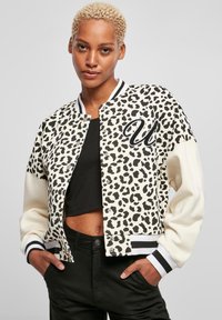 Bomber jakna s uzorkom leoparda s crnim i bijelim detaljima, koja ima preklopnu kopču, rebraste rukave i kontrastne rukave. Ispod je crni skraćeni top.