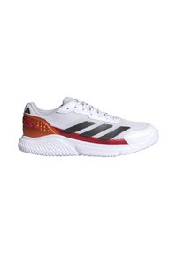 Chaussure de sport blanche avec des rayures noires, des accents rouges et orange au talon, et une semelle blanche texturée orientée vers la droite sur un fond blanc.