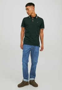 Polo verde con dettagli del colletto bianchi e arancioni a contrasto, abbinato a jeans blu e scarpe marroni; vestibilità standard e stile casual.
