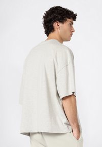 Jonge man met krullend donker haar, gekleed in een losse lichtgrijze t-shirt en lichte broek, staande in profiel met één hand in zijn zak.